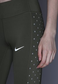 Oliwkowe legginsy sportowe z wysokim stanem, z refleksyjnymi kropeczkami wzdłuż boku i małym białym logo.