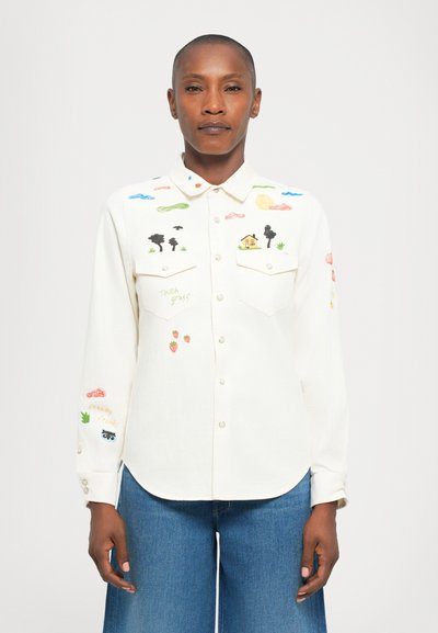 Une personne portant une chemise blanche à boutons ornée d'une broderie colorée sur le thème de la nature et deux poches poitrine à l'avant, associée à un jean bleu.