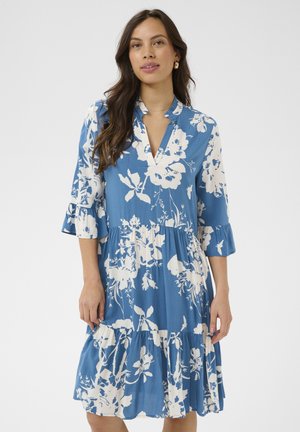 Žena s dlhými tmavými vlasmi, oblečená v modrých šatách po kolená s bielym kvetinovým vzorom a trojštvrťovými rozstrapkanými rukávmi, stojaca na bielom pozadí.