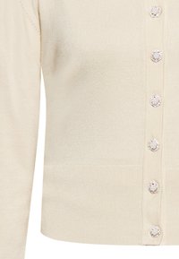 Beige, kötött cardigan, amely passzos szabású, elülső részén hat kerek, drágakőhöz hasonló gombbal, valamint bordázott mandzsettákkal és aljjal.