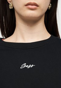 Femme portant des boucles d'oreilles en forme de nœud doré et un t-shirt noir à col rond avec la broderie blanche « Boss » centrée sur la poitrine.