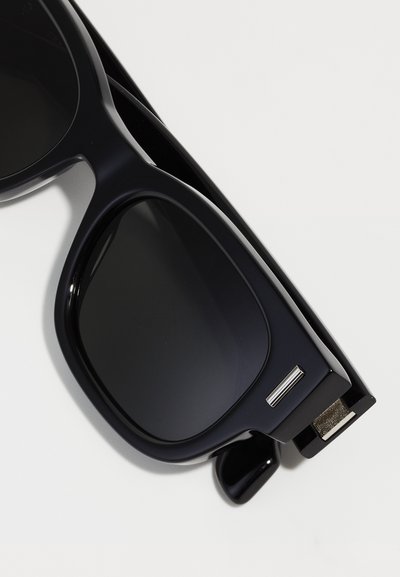Schwarze Kunststoffsonnenbrille mit dunklen Gläsern, rechteckigem Rahmen, dicken Bügeln und metallischen Akzenten an der Seite.
