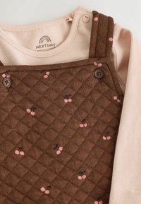 Brązowe pikowane ogrodniczki z różowym wzorem wiśni, zestawione z jasnoróżową bluzką na długi rękaw. Ozdobione guzikami na ramiączkach. Miękka faktura.