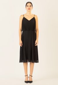 Schwarzes, plissiertes Kleid mit verstellbaren Spaghettiträgern und taillierter Taille. Durchsichtiger Saum, kombiniert mit schwarzen spitzen Schuhen.