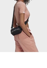 Černá krosbodi taška Adidas se zipovým uzávěrem, s bílým logem, nošena přes světle růžový cropped top a odpovídající tepláky.