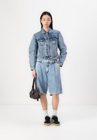 Chaqueta de mezclilla con cierres de botón y dos bolsillos en el pecho, combinada con shorts de mezclilla claros. Los accesorios incluyen un pequeño bolso oscuro y zapatillas negras.