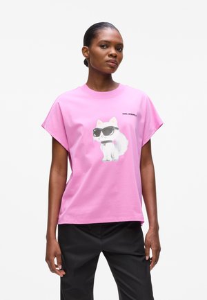 Woman wearing a pink oversized T-shirt with a white cat in sunglasses graphic and black pants, standing against a white background.