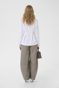 Woman with long wavy hair wearing a white blouse, wide beige pants, white heels, and holding a small brown handbag, standing against a plain background.
