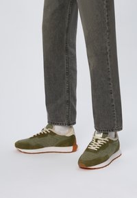 Baskets de sport vertes avec un matériau texturé, des accents en daim, une semelle intermédiaire blanche et une semelle extérieure marron. Associez-les à un pantalon en denim gris et des chaussettes blanches.