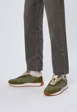 Baskets de sport vertes avec un matériau texturé, des accents en daim, une semelle intermédiaire blanche et une semelle extérieure marron. Associez-les à un pantalon en denim gris et des chaussettes blanches.