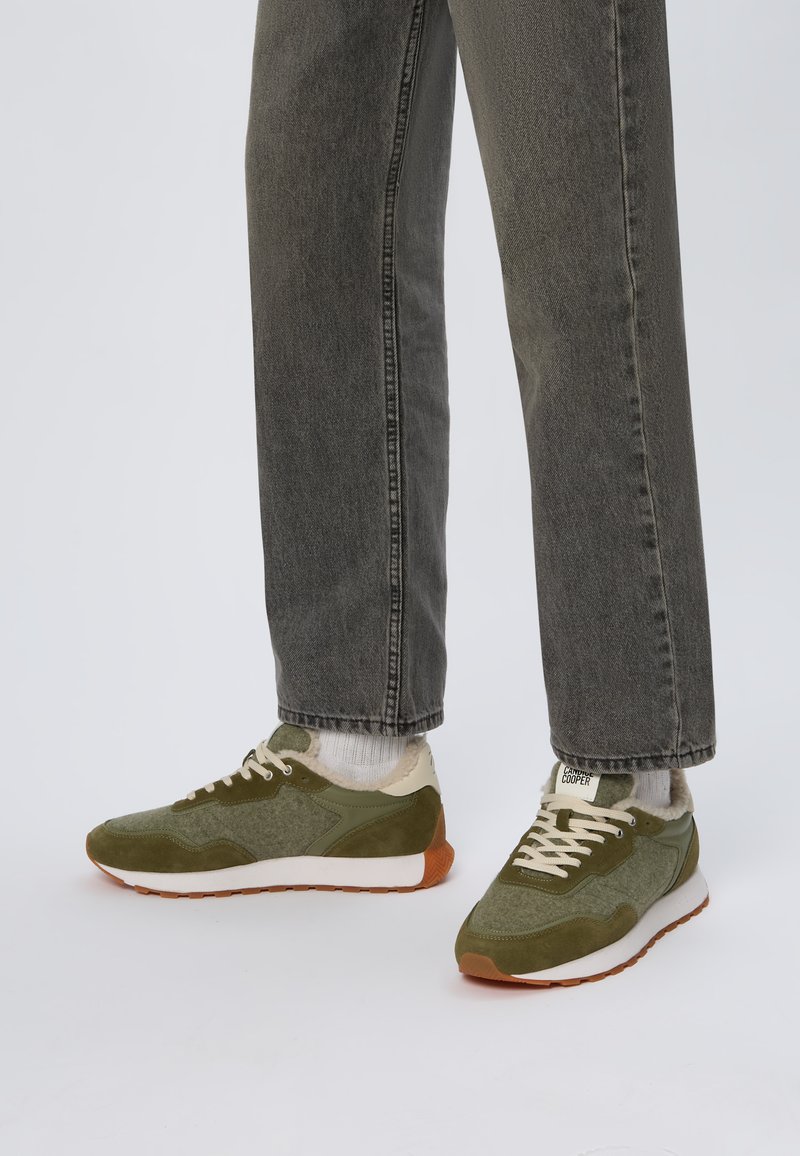 Baskets de sport vertes avec un matériau texturé, des accents en daim, une semelle intermédiaire blanche et une semelle extérieure marron. Associez-les à un pantalon en denim gris et des chaussettes blanches.