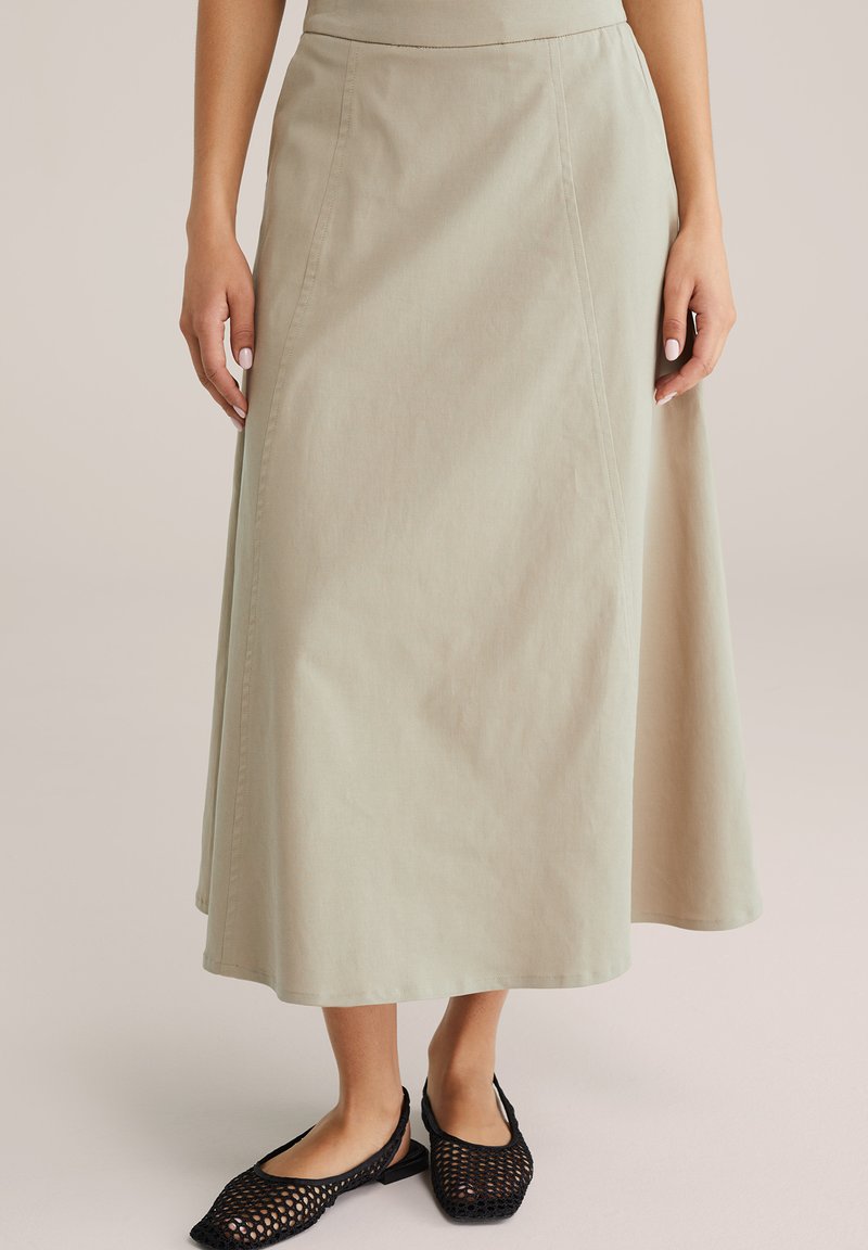 Beige A-formet midi-nederdel båret af en kvinde sammen med sorte flettede flade sko, der viser nedre torso, ben og hænder.