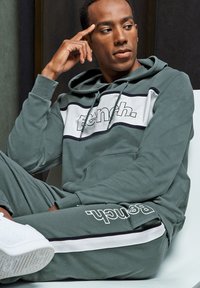 Grüne Kapuzenjacke und passende Jogginghose mit weißen und schwarzen Akzenten, die ein großes Logo tragen. Glattes Material und lockerer Sitz, mit einer Kapuze mit Kordelzug.