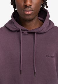 Felpa viola realizzata in morbido tessuto, con cappuccio regolabile tramite cordini e logo ricamato sul petto. Superficie texturizzata e vestibilità rilassata.