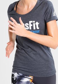 Šedé fitness tričko s logem "CrossFit" v bílé a modré barvě, krátké rukávy, spárované s legínami s vzorem a výbuchy barev.