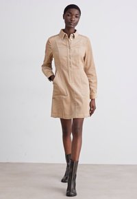 Beige Cordrockkleid mit Knopfleiste, langen Ärmeln, zwei Fronttaschen und tailliertem Schnitt, kombiniert mit kniehohen schwarzen Stiefeln.