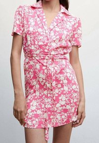 Vestido floral rosa con mangas cortas, cuello, diseño envolvente y cintura ceñida con cinturón. Presenta un patrón floral blanco en toda la prenda. Textura de tela suave.