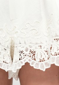 Fustă albă din material textil, adornată cu dantelă florală complicată și modele brodate pe tiv, purtată deasupra genunchilor pe picioare goale.