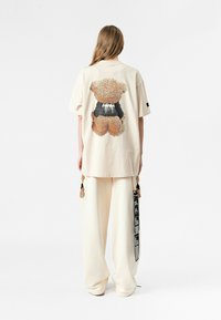 Camiseta beige de gran tamaño con un gráfico texturizado de un oso en la parte de atrás y acentos en negro. Combinada con pantalones de pierna ancha color crema que presentan borlas decorativas.