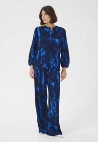 Sininen kuvioitu paita, jossa on pallohihat, yhdistettynä samanlaisiin löysiin housuihin; molemmissa on erilaisia sinisen sävyjä tummalla taustalla.