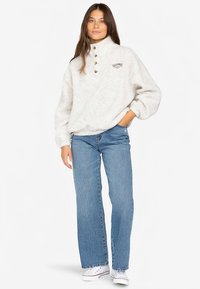 Fleecegenser i off-white med høy krage, tre brune knapper og en liten logo. Kombinert med lyseblå vide jeans og hvite joggesko.