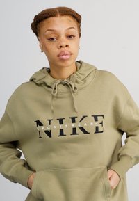 Alyvuogių spalvos megztinis su kapisonu, pagamintas iš minkštos medžiagos, su priekiniais kišenėmis ir veikimo virvele. Juodas "NIKE" logotipas su baltais akcentais priekyje.