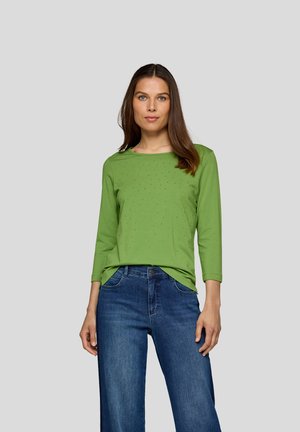 Grünes Langarmshirt mit rundem Ausschnitt, das mit einem strukturierten Muster aus kleinen Verzierungen versehen ist. Kombiniert mit dunkellblauen weit geschnittenen Jeans.