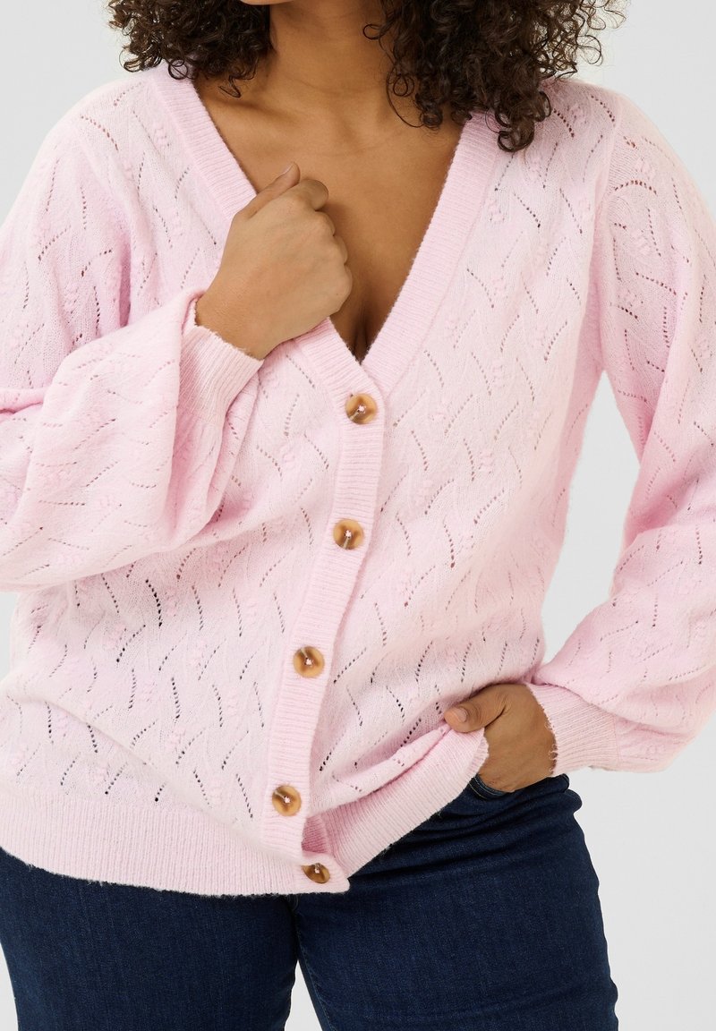 Lys rosa strikket cardigan med V-udskæring, lange ærmer og åben vævning. Har fire brune knapper samt ribbede manchetter og kant.