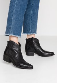 Botas negras hasta el tobillo en cuero texturizado con un patrón de serpiente. Tienen una punta afilada y un tacón bajo de madera. Cierre de cremallera en un lado.