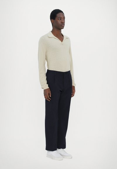 Beige langermet polo med krage, kombinert med mørkeblå bukser og hvite sneakers. Stoffet ser mykt ut med en subtil tekstur.