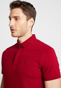 Röd polo skjorta tillverkad av mjukt bomullstyg. Har en krage, knappslå och broderad logotyp på bröstet. Korta ärmar.