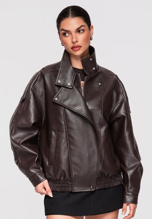 Kobieta o ciemnych włosach związanych w niskiego koka, ubrana w oversize ciemnobrązową skórzaną kurtkę z elastycznym ściągaczem i czarną spódnicę, patrzy w bok.