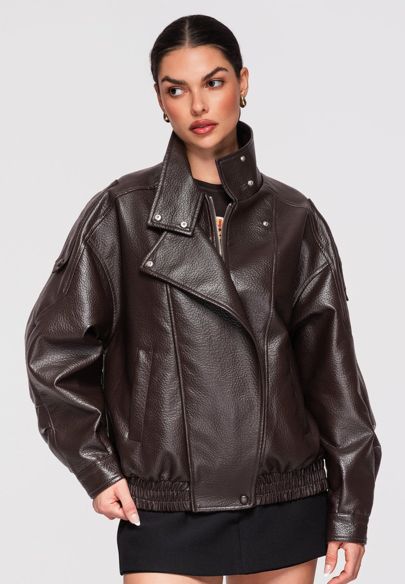 Mujer con cabello oscuro recogido en un moño bajo, vistiendo una chaqueta de cuero marrón oscuro de gran tamaño con dobladillo elástico y falda negra, mirando hacia un lado.