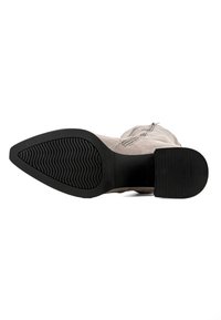 Cizmă de glezna din suede bej cu o talpă de cauciuc neagră, având o formă curbată și un model texturat al benzii de rulare. Fermoarul lateral este vizibil pentru închidere.