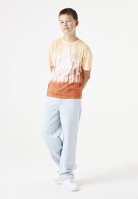 Camiseta de algodón de manga corta en color amarillo y naranja con un diseño tie-dye, que presenta un logo sutil. Se combina con pantalones de mezclilla azul claro y zapatillas blancas.