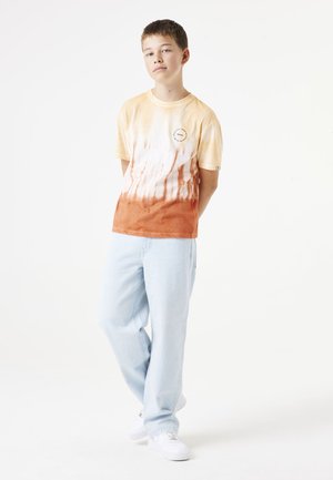 Camiseta de algodón de manga corta en color amarillo y naranja con un diseño tie-dye, que presenta un logo sutil. Se combina con pantalones de mezclilla azul claro y zapatillas blancas.