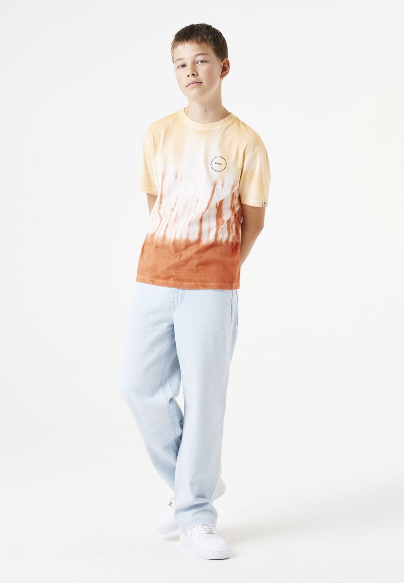 Camiseta de algodón de manga corta en color amarillo y naranja con un diseño tie-dye, que presenta un logo sutil. Se combina con pantalones de mezclilla azul claro y zapatillas blancas.