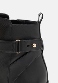Gros plan sur une botte cheville en cuir noir avec une bride et des clous métalliques dorés sur un fond clair.