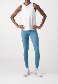 Top deportivo blanco sin mangas con cuello redondo, combinado con leggings de color azul verdoso. La modelo lleva zapatillas blancas; la tela parece ligera y suave.