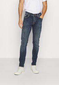 Jeans en denim bleu avec une coupe slim, un design à cinq poches et des coutures contrastées. Portés avec un t-shirt blanc et des baskets de couleur claire.