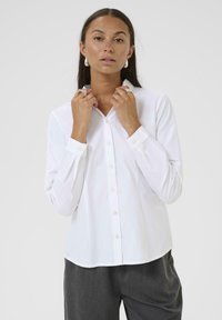 Chemise blanche à boutons, avec un col, des manches longues et une texture lisse. Présente un ourlet arrondi et des boutons assortis.