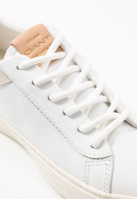 White leather sneaker with beige accents, flat white laces, and a tan leather tag. Features textured surface and rubber sole.