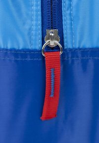 Dettaglio della cerniera di una borsa blu con un pull tab argento, un'etichetta in nylon rossa sulla cerniera e un pannello in tessuto blu a contrasto sopra.