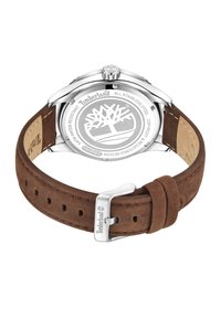 Reloj con correa de ante marrón y caja de acero inoxidable plateado, con logotipo de la marca visible y patrón de árbol grabado en la parte posterior. Resistente al agua.
