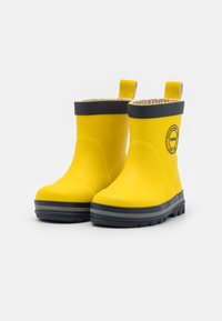 Une paire de bottes de pluie jaunes imperméables pour enfants avec des bordures noires, des poignées de tirage et des semelles texturées sur un fond blanc.