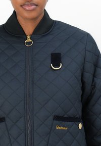 Navy quilted jacket with ribbed collar, gold hardware accents, front pockets, and embroidered "Barbour" logo. Circular zipper pull detail.