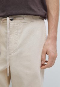 Beige corduroy trousers with an elastic waistband and drawstring. Features a front pocket and a black button. Texture is ribbed.