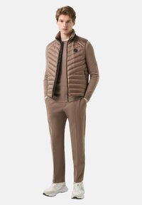 Braune Steppweste mit Reißverschluss über einem hellbraunen Pullover, kombiniert mit passenden braunen Hosen und weißen Sneakern.