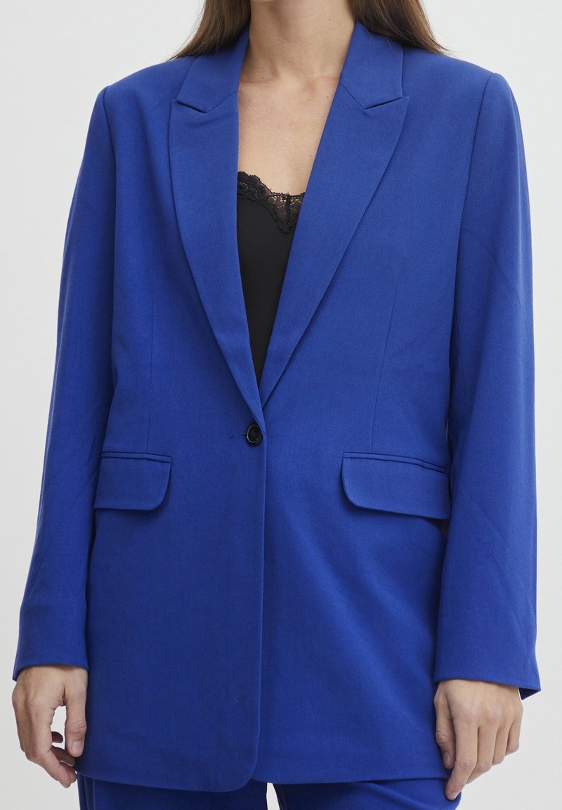 Femme portant un blazer bleu royal avec un seul bouton et des poches à rabat sur un haut noir orné de dentelle, visible du cou jusqu'à mi-cuisse.