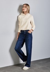 Felpa beige chiaro con cappuccio, abbinata a jeans di denim a gamba larga blu scuro e sneaker bianche, su uno sfondo neutro.